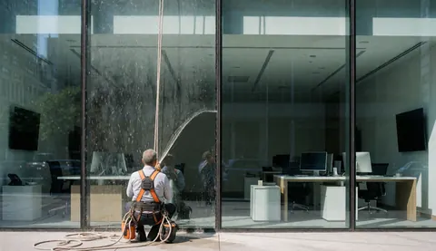 Worker in harness cleaning large office window with visible interior desks and computers