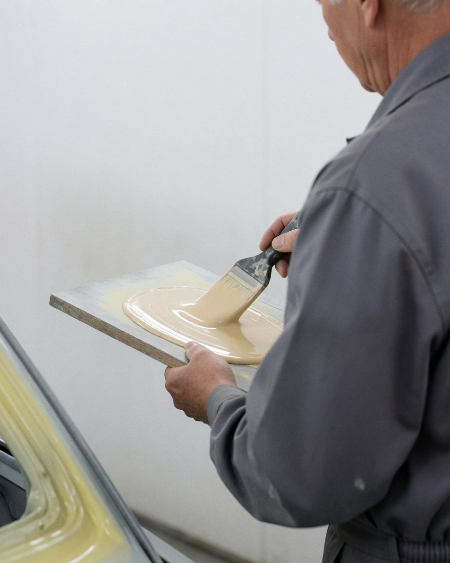 Technician mixing body filler on a panel with a brush in a white-walled paint shop