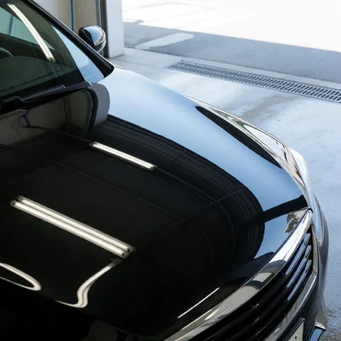 Polished black car hood reflecting fluorescent lights above a concrete floor with a drainage grate in a car wash area