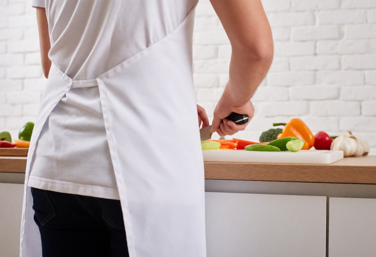A person wearing a white apron is chopping fresh vegetables in a kitchen with a white brick wall background.