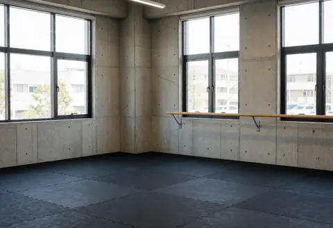 Empty modern dance studio featuring a wooden barre, concrete walls, large industrial windows, and black floor mats suitable for dance classes.
