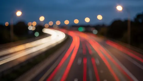 Red and white vehicle light streaks on an elevated road at night with soft blue and orange bokeh, ideal as a text background.