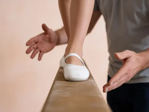 Student feet in white shoes on wooden beam with coach hands ready to assist during personalized gymnastics training