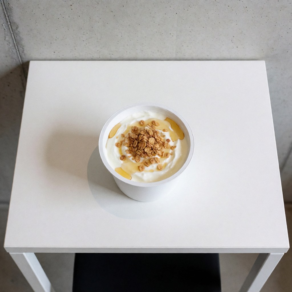 Greek yogurt topped with honey and granola in a white cup on a white table in an apartment hotel with concrete wall, overhead view