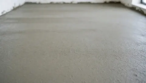 A freshly poured concrete floor with white walls and a window, serving as a neutral background for tennis court installation text.