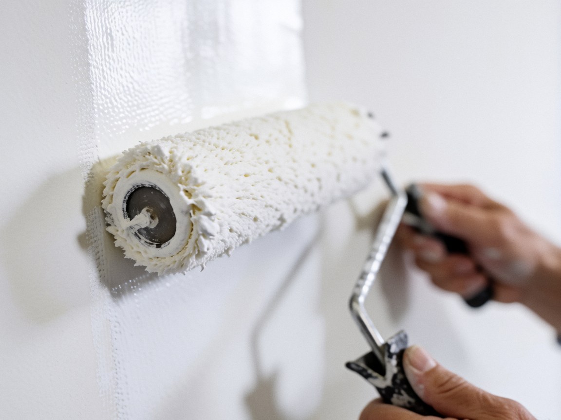 Close-up of hands using a white roller to apply fresh white paint to a wall, showing fiber texture and bright light for wall repairers.