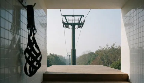 A black zipline harness hangs on a white tiled wall with a wooden tower and platform in the background for adventure parks.