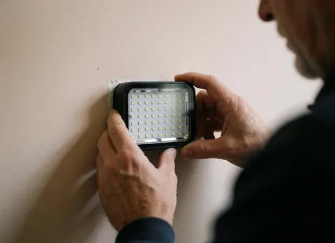 Close-up of hands mounting an LED spotlight with visible diodes on a beige wall, showing tennis court lighting installation details.