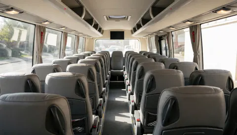 Interior view of a tour bus featuring rows of gray seats, a central aisle, and natural light streaming through large windows.