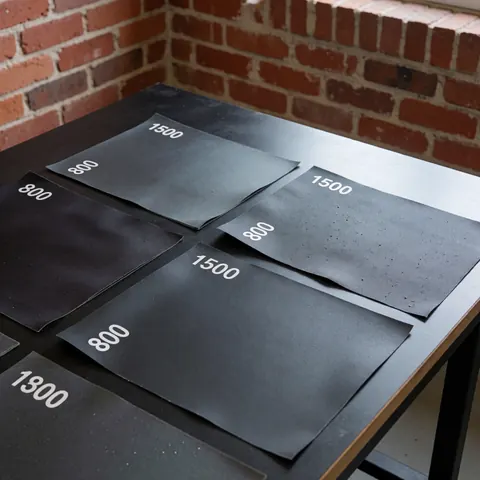 Close-up of automotive sandpaper sheets with 800 and 1500 grit textures resting on a dark work surface against a red brick wall for auto body repair