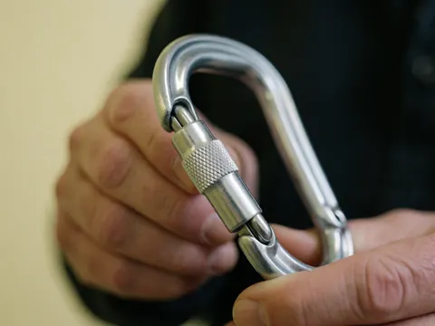 Close-up of an adult hand holding a shiny metal climbing carabiner against a pale yellow wall, highlighting the safety mechanism detail.