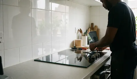 A technician installs a glass cooktop in a modern kitchen with white tiled walls and visible tools.