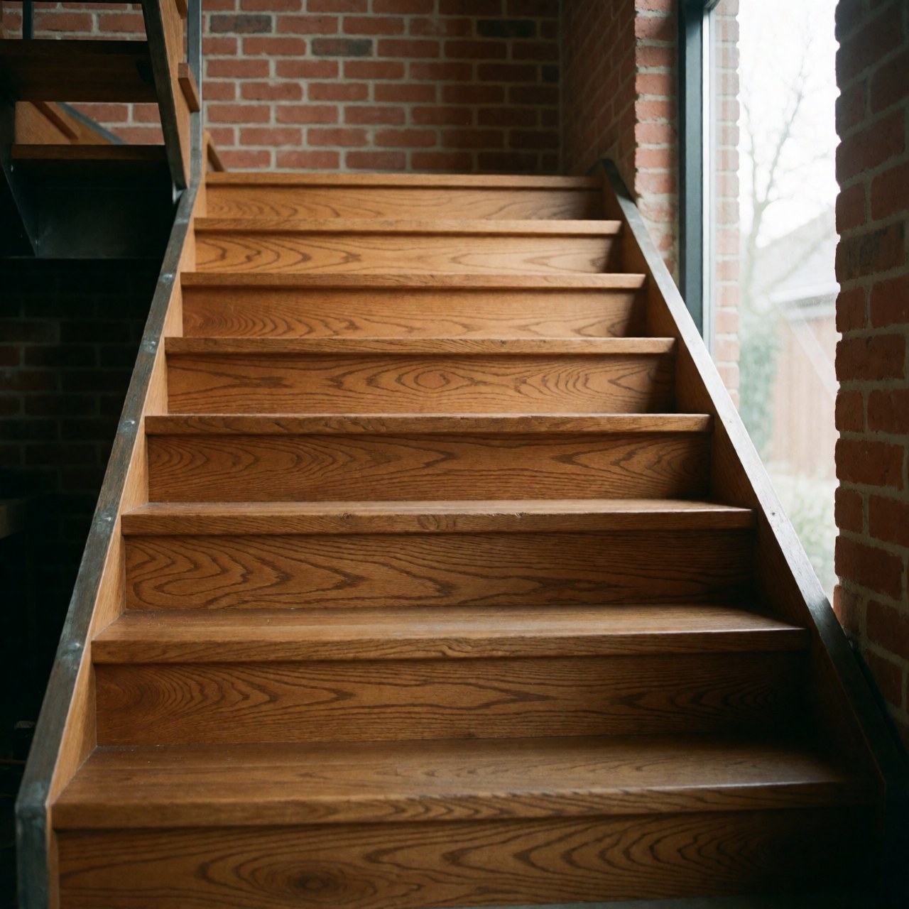 Oak wood staircase with metal structure in a hallway with brick walls, illuminated by natural light.