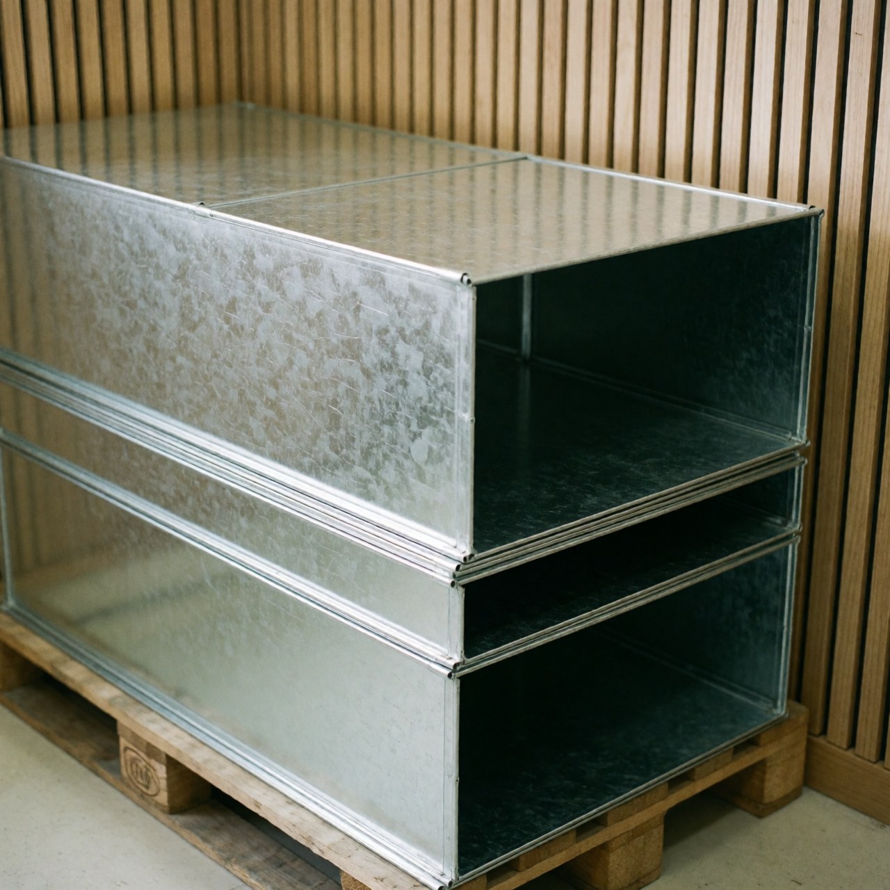 Stacked galvanized metal ducts on a wooden pallet with slat walls and natural lighting for metal stamping business