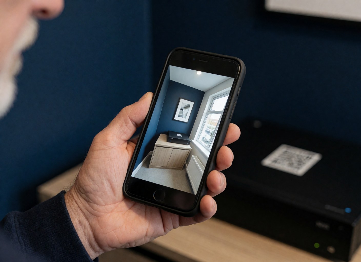 Hand holding a smartphone displaying a security camera feed in a room with navy blue walls and an NVR unit with a QR code