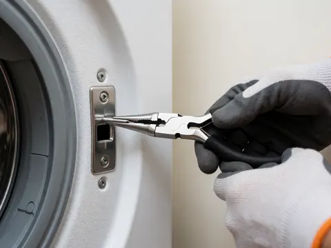 Gloved hand inserting a metal pin into the textured white interior of a washing machine door using pliers