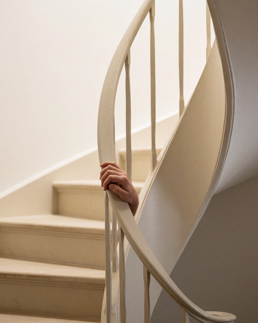 Close-up of a hand touching the railing of a spiral staircase against a white wall with warm lighting highlighting the finish details