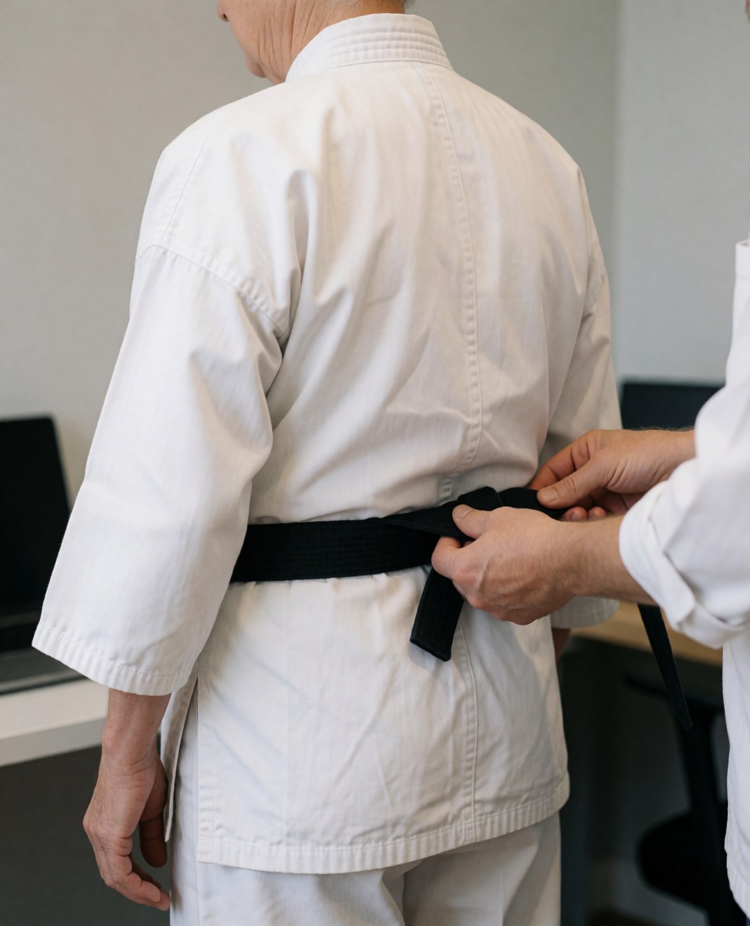 Karate instructor tying a black belt for a student in a dojo with light gray walls