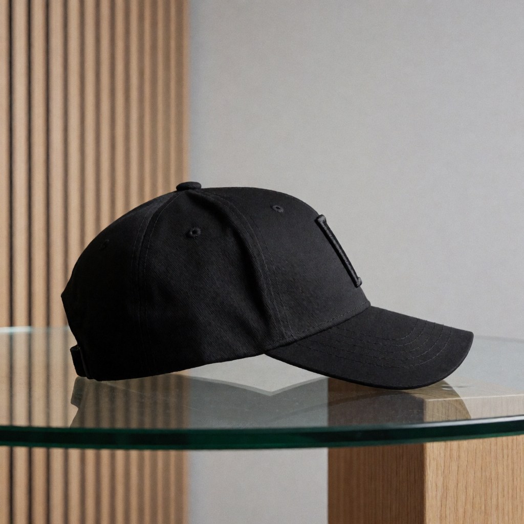 Black embroidered baseball cap resting on a clear glass table with a gray background and light wood panels, suitable for motorsport merchandise catalogs