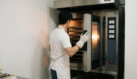 A baker wearing gloves and a white apron opens an industrial oven with a thermometer against white walls, leaving space for text on the left.