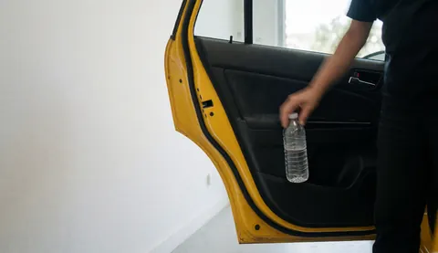 A hand reaches into the door pocket of a yellow taxi to grab a water bottle, with a white wall and copy space on the left