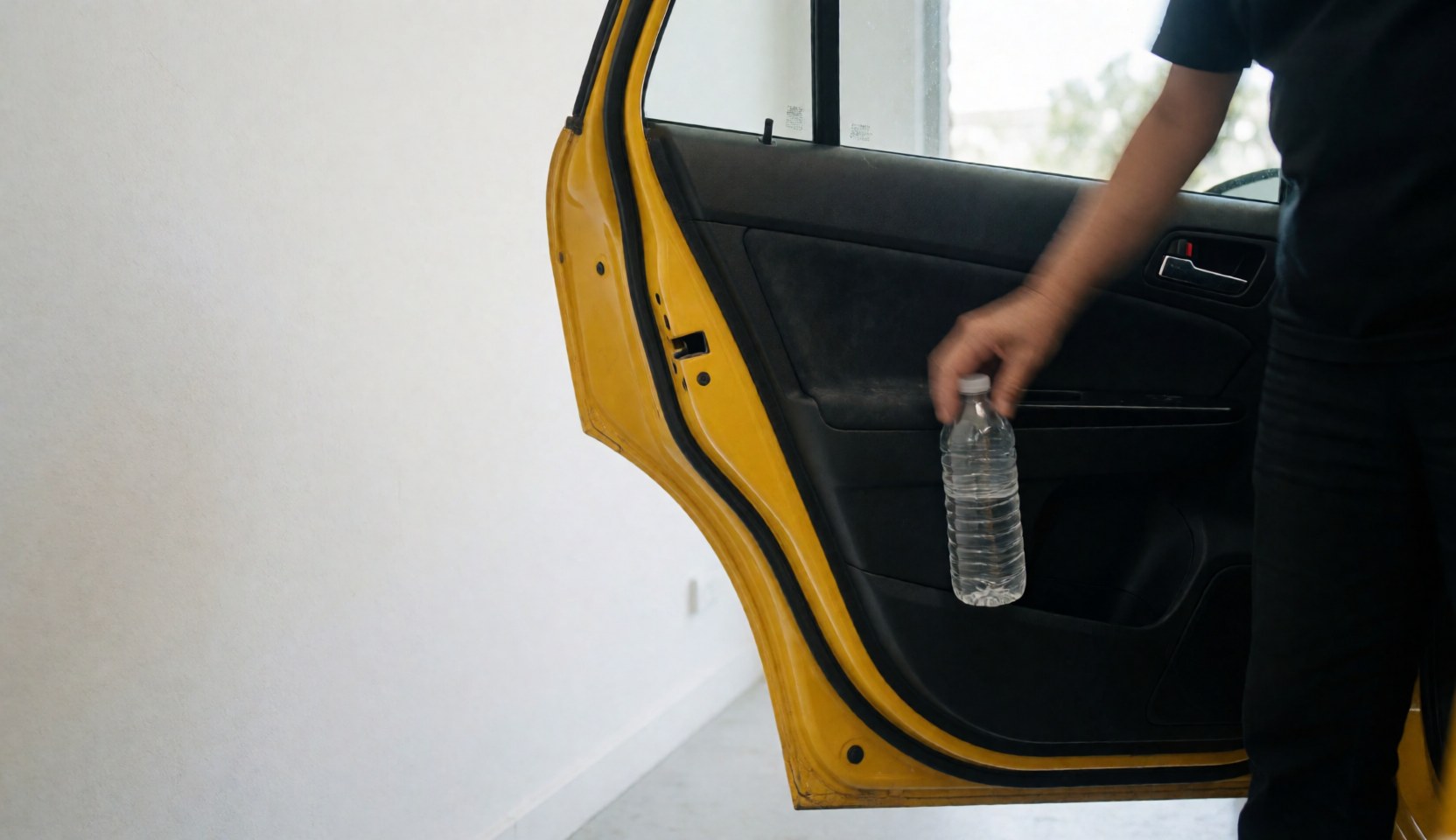 A hand reaches into the door pocket of a yellow taxi to grab a water bottle, with a white wall and copy space on the left