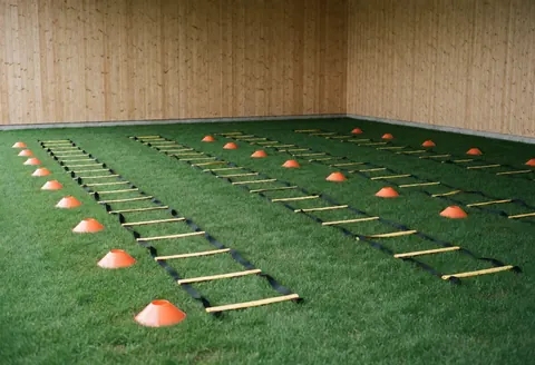 Soccer training area featuring agility ladders and orange cones on artificial turf with wooden walls, no people present