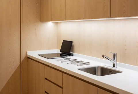 White countertop featuring a stainless steel sink and various tools, set against light wood panels for a clean homeopathy clinic interior.