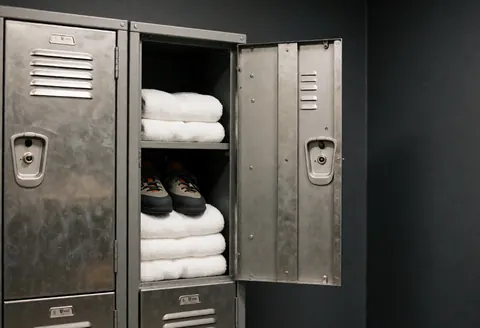 Open metal locker containing climbing shoes and white towels against a charcoal gray wall in a climbing gym locker room