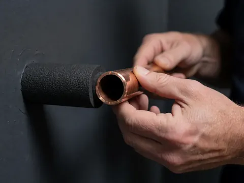 Close-up of a hand adjusting a copper pipe with black insulation against a dark gray wall, showing heating system installation details.