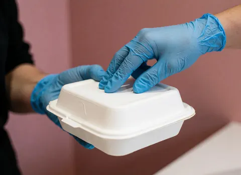 Hands wearing nitrile gloves sealing a white food container against a pink wall to show hygiene in a poultry shop