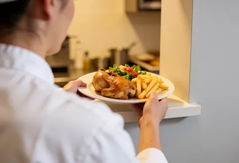 A cook serves a plate of roasted chicken, french fries, and green salad on a white counter in a professional kitchen.