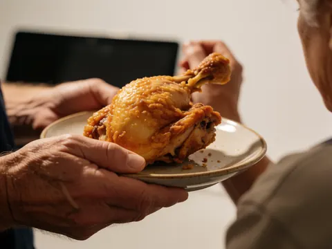 Adult hands holding a fried chicken quarter on a plate against a white wall with warm lighting