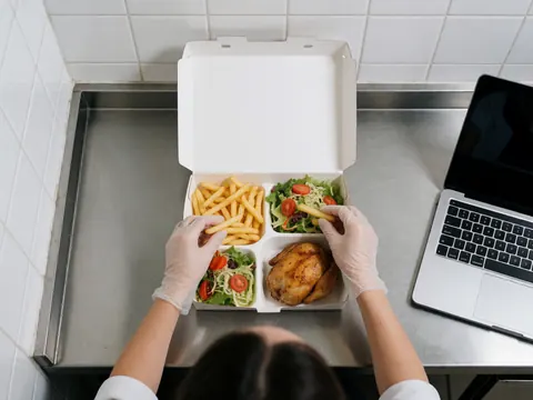 Hands wearing gloves place fried chicken, fries, and salad into a takeout box on a stainless steel counter in a tiled kitchen.