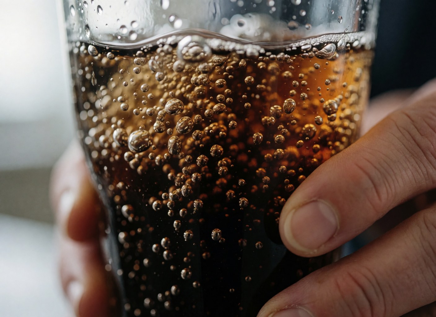 Close-up of carbonated bubbles in a dark beverage inside a clear glass, held by a hand with natural light and reflections on the glass surface