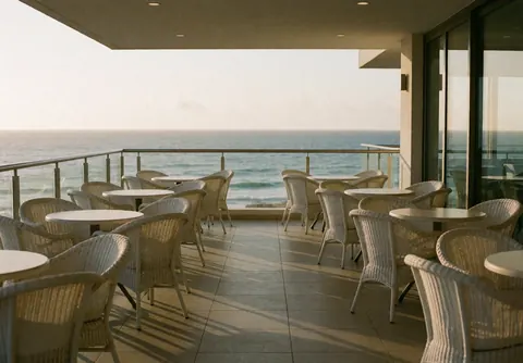 Outdoor terrace of a cevicheria featuring round tables and wicker chairs overlooking the sea, with tiled flooring and a steel railing.