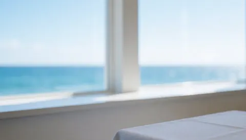 White tablecloth on a table near a window overlooking the sea with white walls, ideal for coastal restaurant advertising background