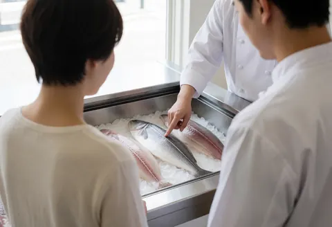 Chef in white uniform points to fresh fish on ice in a stainless steel container while serving a customer in a fresh food establishment.