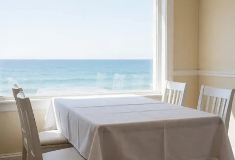 Empty dining table with white tablecloth and chairs near a window overlooking the ocean, featuring beige walls and natural light.