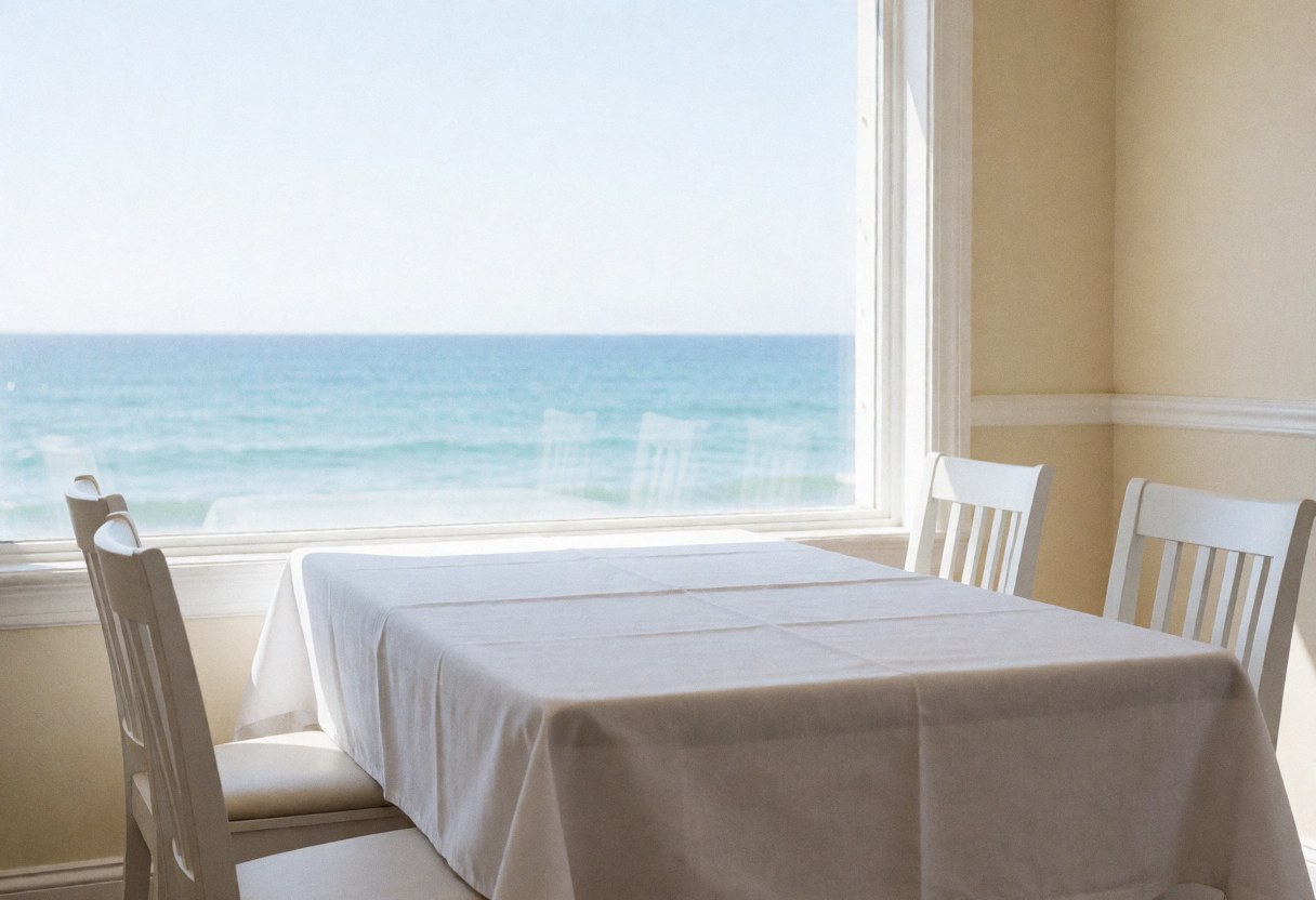 Empty dining table with white tablecloth and chairs near a window overlooking the ocean, featuring beige walls and natural light.