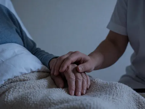 Close-up of a caregiver's hand gently holding the hand of an elderly person under a white blanket in a quiet setting.