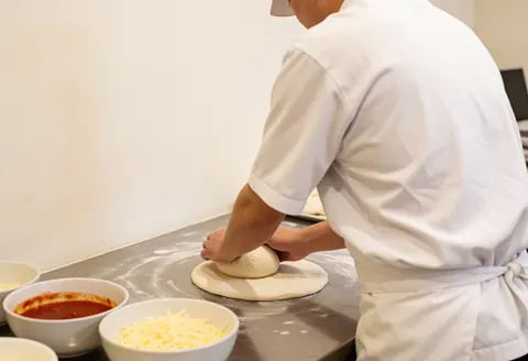 Chef stretching pizza dough on a stainless steel counter with bowls of sauce and cheese nearby in a clean kitchen.