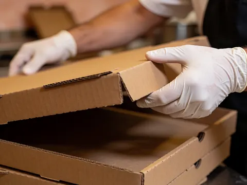 Gloved hands seal a cardboard pizza box in a kitchen with terracotta walls, preparing it for delivery.