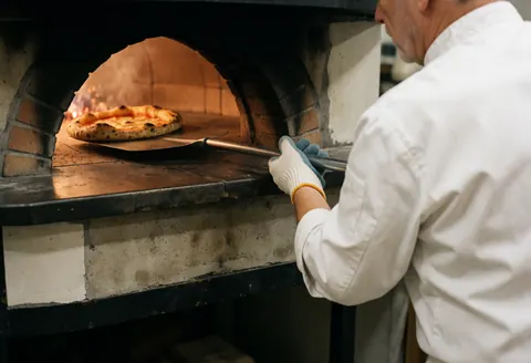 A chef wearing protective gloves uses a peel to take a freshly baked pizza out of a hot brick oven with visible flames.