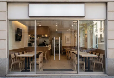 Restaurant entrance featuring tables and chairs with a person working in the kitchen under warm modern lighting