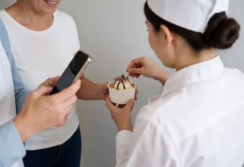 Server adjusting chocolate and nut topping on ice cream while a customer takes a photo in a light gray walled shop