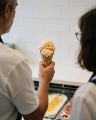 A customer receives a waffle cone filled with ice cream at a shop with white tiled walls.