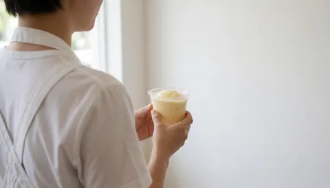 Person serving ice cream in a cup against a white wall with natural light and space for text