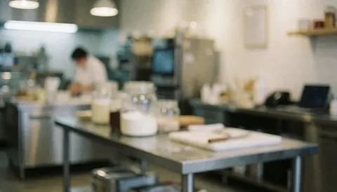 Kitchen background for an ice cream shop featuring stainless steel tables, glass pitchers, and a person working in the background under soft lighting