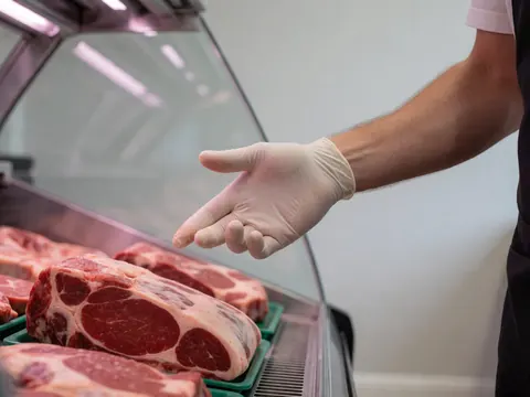 A gloved hand points to a cut of meat inside a refrigerated display case against a light gray wall
